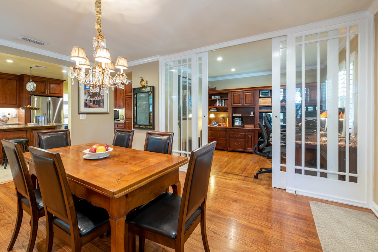 A cozy dining room with a wooden table, leather chairs, and a chandelier, connected to an office space.